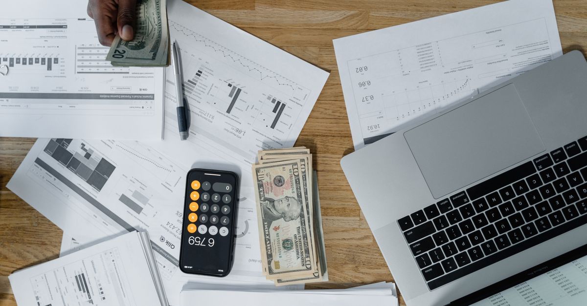 A desk with a laptop, paperwork, a calculator and cash
