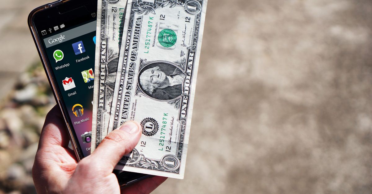 A hand holding a phone showing a banking app, next to a dollar bill